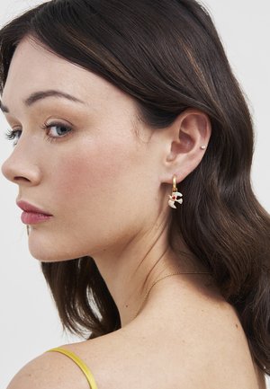 Približna snimka žene s tamnosmeđom kosom koja nosi zlatne naušnice u obliku obruča s privjescima bijelih golubica s crvenim detaljima.