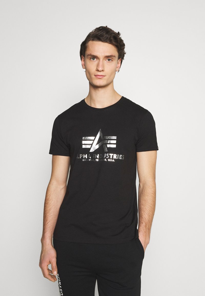 Tricou din bumbac negru cu guler rotund, având un logo metalic de culoare argintie și elemente de design pe față, asortat cu o pereche de pantaloni de trening închiși la culoare.