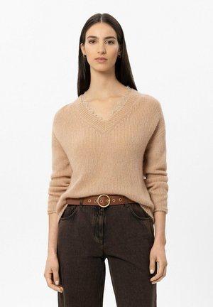 Woman with long dark hair wearing a beige knit sweater with a V-neck and dark high-waisted jeans with a brown belt with a round buckle.