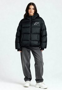 Doudoune noire avec capuche, ornée d'un cœur blanc et d'une flèche sur la poitrine, associée à un jean gris ample et des baskets blanches.