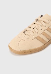 Beige mockasinsneaker med texturerad yta, med tre bruna ränder, snörning framtill och gummisula med sydd kant.