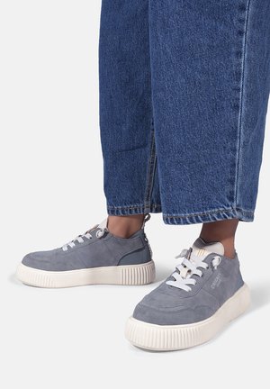 Persona che indossa jeans blu a gamba larga e sneakers platform in suede grigio con lacci bianchi e suole bianche spesse su sfondo bianco.