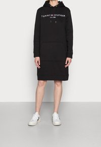 Svart hoodieklänning i mjukt material, med en framficka, dragsko-huva och logotext vid bröstet. Stylad med vita sneakers.