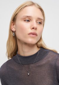 Blond kvinna iklädd en grå transparent överdel, silverhjärta hängande halsband och matchande silver hoopörhängen med små hjärtan, ser åt vänster.