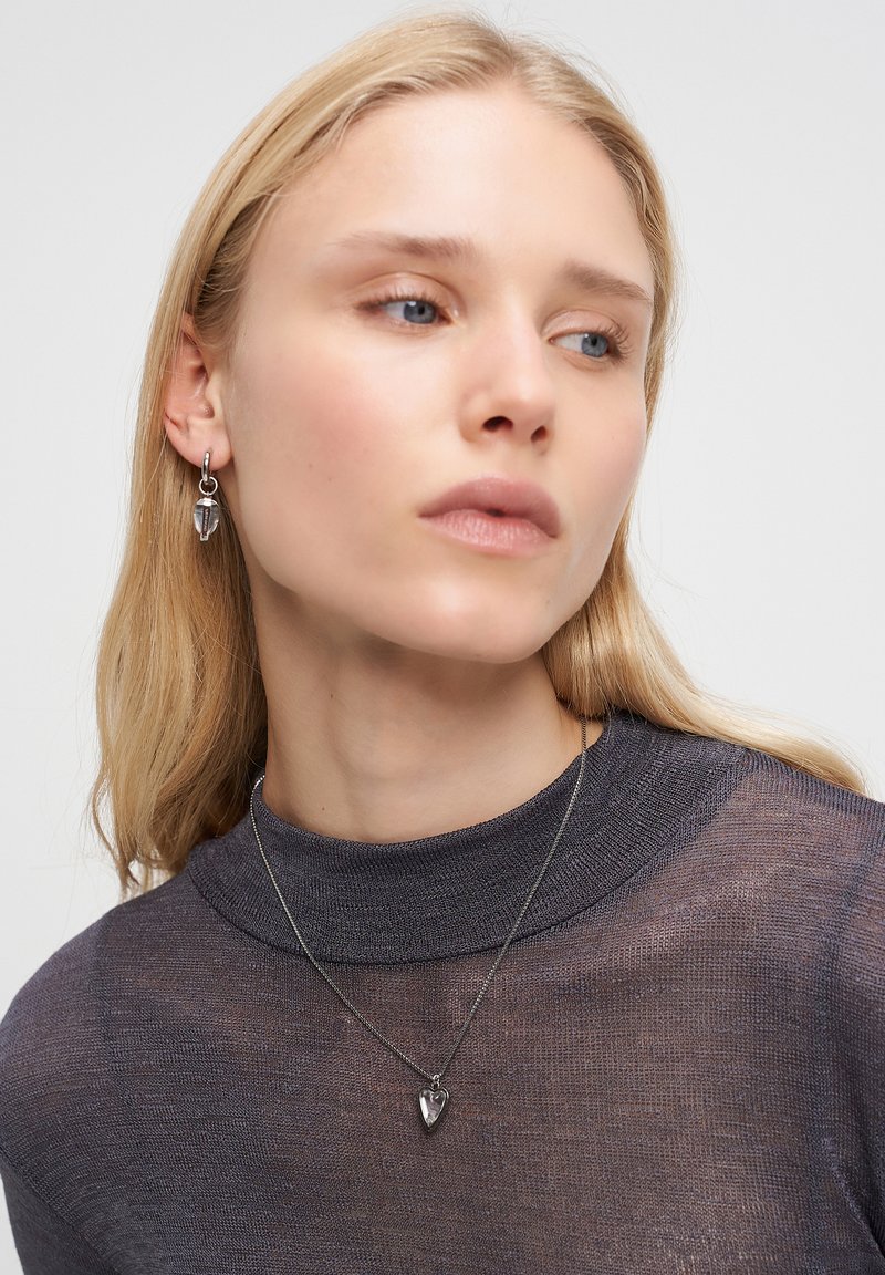 Blond kvinna iklädd en grå transparent överdel, silverhjärta hängande halsband och matchande silver hoopörhängen med små hjärtan, ser åt vänster.