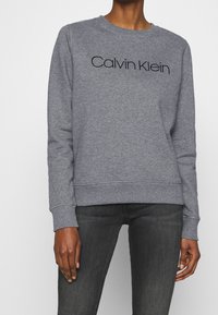 Sudadera gris con cuello redondo y mangas largas. Presenta el logo en negro "Calvin Klein" en la parte frontal. Está hecha de una tela suave y texturizada.