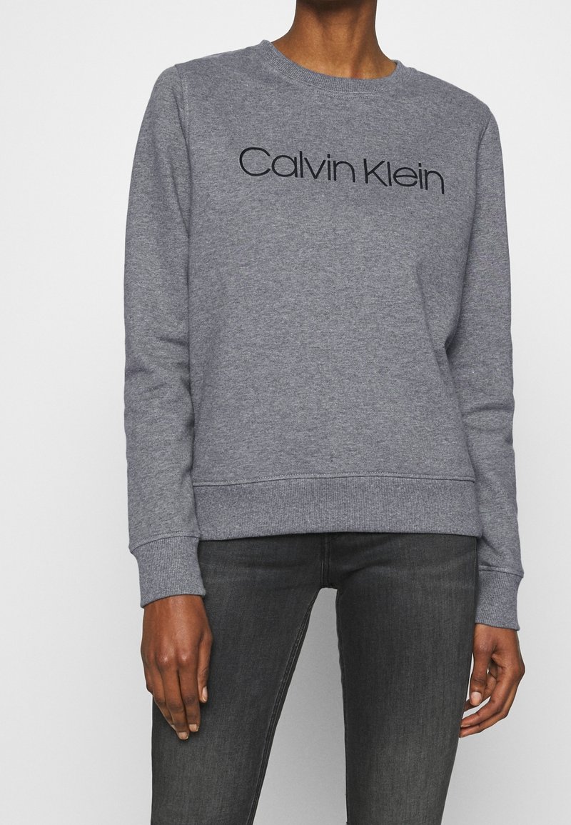 Sudadera gris con cuello redondo y mangas largas. Presenta el logo en negro "Calvin Klein" en la parte frontal. Está hecha de una tela suave y texturizada.