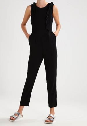 Frau trägt einen ärmellosen schwarzen Jumpsuit mit Rüschen auf der Vorderseite, Hände in den Taschen und silberne Sandalen mit Schnalle vor weißem Hintergrund.