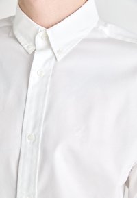 Chemise en coton blanc à boutonner avec un col classique, ornée d'une broderie de logo discrète et de boutons unis, texture lisse et coupe ajustée.