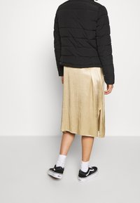Giacca a vento nera con texture trapuntata, abbinata a una gonna lucida dorata di lunghezza media con uno spacco laterale, indossata con scarpe da ginnastica nere.