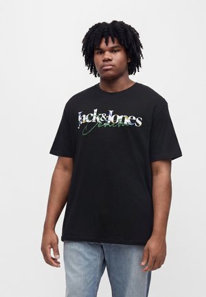 Mladý muž s kučeravými vlasmi, oblečený v čiernom tričku Jack & Jones Jeans a svetlomodrých džínsoch, stojí na jednofarebnom pozadí.