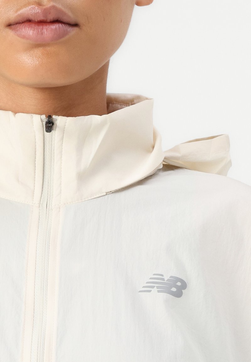Veste en crème légère avec un col haut, fermeture éclair à l'avant et texture douce. Présente un logo gris subtil sur la poitrine et des accents à nouer à l'arrière.