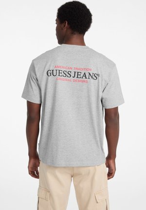 Grå bomulds t-shirt med "GUESS JEANS" i sort og rød tekst på ryggen, rund halsudskæring, korte ærmer og en afslappet pasform.
