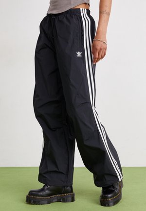 Personne portant un pantalon de survêtement Adidas noir avec des bandes blanches et des bottes épaisses noires, debout sur un sol vert devant un mur blanc.