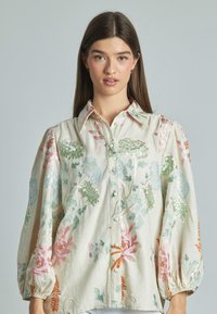 Blusa ricamata con colletto, maniche lunghe e bottoni. Presenta motivi floreali in verde, blu, rosa e arancione su uno sfondo beige.