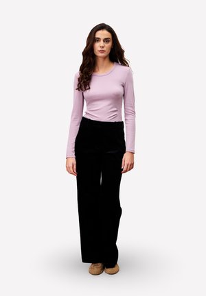 Femme aux longs cheveux ondulés, portant un haut à manches longues violet clair, un pantalon large noir et des chaussures beiges, se tenant devant un fond uni.