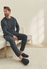 Ensemble de vêtements d'intérieur en maille gaufrée noire avec manches longues et pantalon, accompagné de chaussons noirs rembourrés, assis sur un tapis clair.