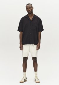 Svart kortärmad skjorta med knappar och bröstficka, matchad med vita texturerade shorts och beige sneakers med svarta ränder.