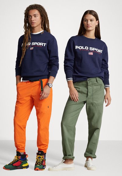 Două persoane stau una lângă alta. Una poartă un hanorac bleumarin cu text alb "POLO SPORT", pantaloni portocalii și adidași multicolorați. Cealaltă are un hanorac bleumarin, pantaloni verzi și pantofi albi.