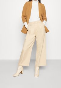 Pantaloni beige a gamba larga con pieghe frontali, abbinati a un dolcevita bianco aderente e a un blazer marrone. Stivali alla caviglia con tacco grosso color crema.