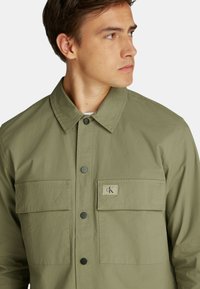 Giacca button-up verde oliva, con colletto a punta, due tasche sul petto e una piccola patch con logo. Realizzata in tessuto strutturato con un design uniforme.