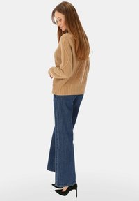 Beżowy dziergany sweter z warkoczowym wzorem, zestawiony z ciemnymi, szerokimi dżinsami i czarnymi czółenkami na szpic, modelka zwrócona bokiem.