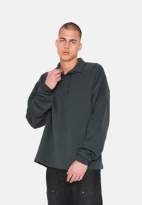 Pullover grigio scuro con colletto, maniche lunghe, vestibilità rilassata e logo ricamato sul pattino. Tessuto morbido con polsini a coste.