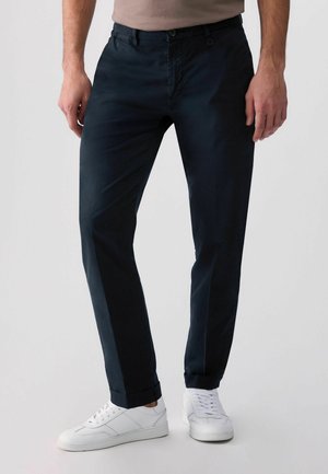 Bărbat purtând pantaloni slim fit bleumarin închis și adidași albi, stând pe un fundal deschis.