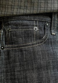 Τσέπη από σκούρο τζιν denim με ορατές ραφές και μεταλλικά καρφιά, συνδυασμένη με γκρι υφαντό ύφασμα με καρό, που έχει μπει μέσα στη μέση του παντελονιού.