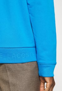 Sudadera azul brillante con puños y dobladillo acanalados, con un logo texturizado en la parte inferior. Combinada con pantalones marrones texturizados.