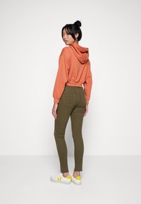 Un sweat à capuche court couleur rouille-orangé, associé à un pantalon moulant vert olive et des baskets blanches avec des accents jaunes. Textures lisses dans l'ensemble.