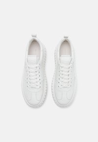 Paire de baskets en cuir blanc avec semelles en caoutchouc épaisses et lacets, vue de dessus sur un fond clair uni.