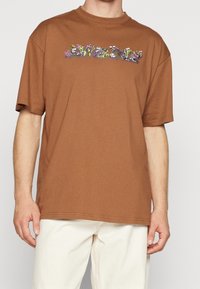 Brun bomullströja med korta ärmar, prydd med en färgglad blommig broderi över bröstet. Rakt snitt med rund halsringning.