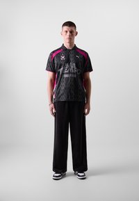 Sort og pink kortærmet trøje med et dækspor-mønster og broderet logo, parret med løse sorte bukser og sort-hvide sneakers.