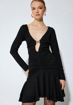 Black dress with long sleeves, deep V-neck, ruched detailing, fitted bodice, flared skirt, and gold-tone hardware accent at the neckline.