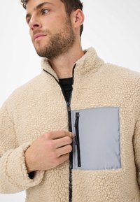 Giacca in fleece beige con tessuto testurizzato, colletto alto, zip frontale e tasca grigia con zip nera. Aspetto morbido e accogliente.