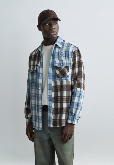 Homme portant une chemise à carreaux bleue et marron par-dessus un t-shirt beige, un jean délavé et une casquette de baseball marron foncé, regardant sur le côté.