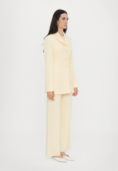 Light yellow tailored suit featuring a long-sleeve blazer with three buttons and wide-leg trousers, made of smooth fabric. Pointed mules in white.