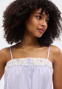 Γυναίκα με σγουρά μαλλιά που φοράει αμάνικο τοπ ανοιχτού μοβ χρώματος με λουλουδένιες και ριγέ λεπτομέρειες, κοιτάζει ελαφρώς πλάγια και χαμογελά απαλά.