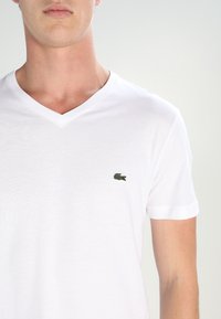 T-shirt en coton blanc à col en V, avec un petit logo crocodile vert brodé sur le côté gauche de la poitrine. Manches courtes et texture douce.
