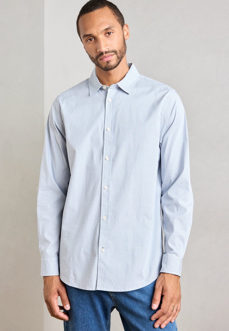 Uomo in camicia a maniche lunghe blu chiaro a scacchi e jeans blu, in piedi contro uno sfondo chiaro e uniforme.