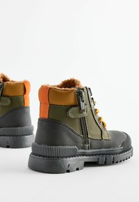 Bottes vertes et grises avec des accents orange, doublure en fourrure, fermeture éclair distincte et semelles en caoutchouc texturé pour une meilleure adhérence. Design montante avec une structure renforcée.