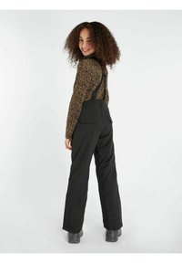 Pantalones de trabajo negros con tirantes ajustables. La tela parece duradera. Combinados con una blusa de manga larga de estampado de leopardo marrón, llevada con zapatos grises.
