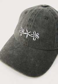 Temno zelena kapa iz mehkega blaga, z značilnimi tremi belimi vezenimi cvetovi spredaj. Ukrivljen rob, teksturirana površina in nastavljiv trak.