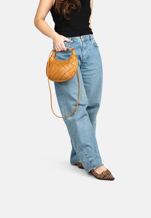 Vrouw draagt lichtblauwe spijkerbroek, zwart mouwloos topje, panterprint ballerina's, houdt een bruine geweven handtas met lange riem vast.