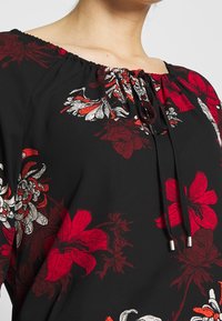 Blusa negra con estampado floral en rojo y blanco, con escote elástico y cordón ajustable con detalles metálicos.