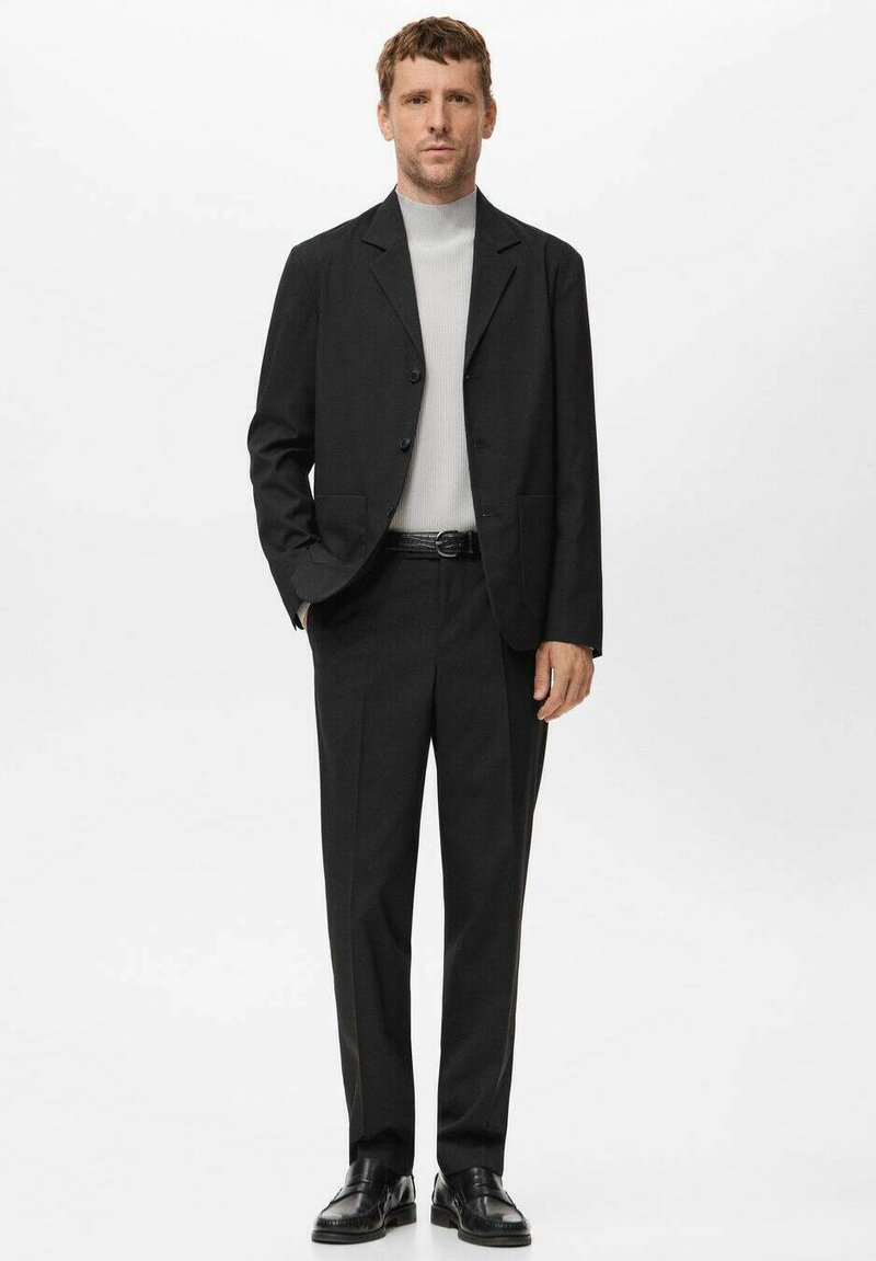 Mann steht in einem schwarzen Anzug, hellgrauem Rollkragenpullover, schwarzem Gürtel und schwarzen Lederschuhen vor weißem Hintergrund.