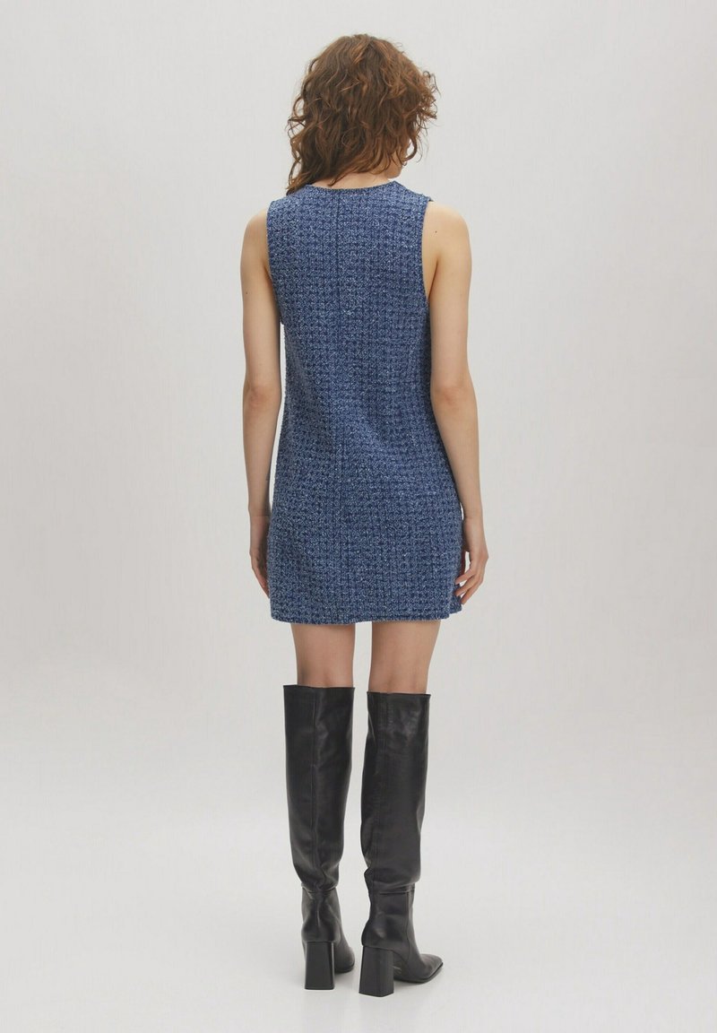 Femme debout, dos face à la caméra, portant une mini-robe bleue texturée sans manches et des bottes noires à talons montant jusqu'aux genoux.