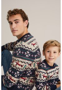 Stickade tröjor i beige med marinblå detaljer, med geometriska och julinspirerade mönster av träd och renar. Denimjeans som bärs av vuxen.