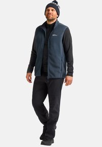 Dunkelblauer Fleece-Weste mit hohem Kragen, Frontreißverschluss und seitlichen Taschen, getragen über einem schwarzen Langarmhemd und kombiniert mit dunklen Hosen und Stiefeln.